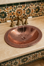 A hammered copper bathroom sink with an ornate brass faucet, installed on a beige stone countertop against a decorative mosaic tile backsplash.