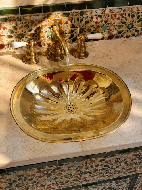 A polished brass oval bathroom sink with a matching three-hole faucet and running water, set into a beige stone countertop against a decorative mosaic tile backsplash.