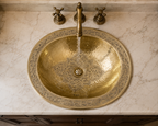 A hammered brass bathroom sink with intricate engraved detailing around the rim, set into a white marble countertop with a matching brass faucet and handles.