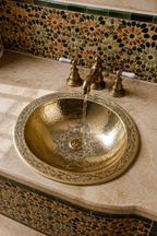 A hammered brass bathroom sink with a matching faucet, set into a beige stone countertop against a decorative mosaic tile backsplash.