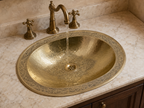An oval hammered brass bathroom sink with an ornate decorative rim, set into a beige marble countertop with a matching brass faucet running water into it.