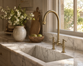 Handcrafted aged brass bridge kitchen faucet installed on a white marble farmhouse sink.