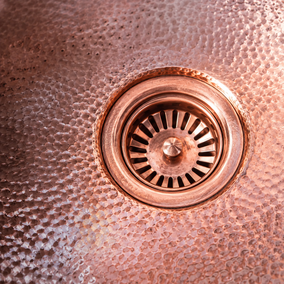 Close-up of a hammered copper sink drain with a matching copper strainer basket and surrounding textured copper basin.