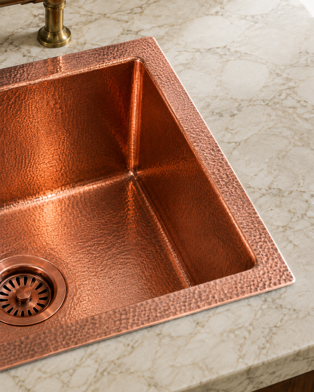 A hammered copper single-basin sink set into a white marble countertop, with a matching copper drain and part of a brass faucet visible.