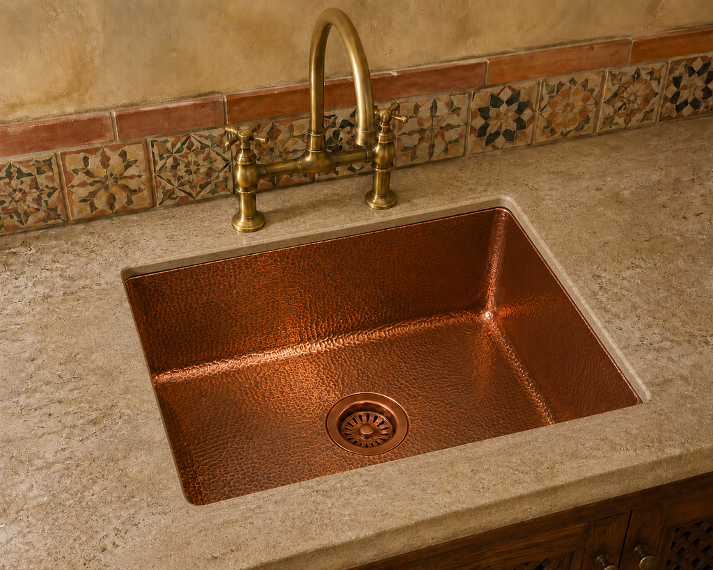 A hammered copper farmhouse sink set into a speckled beige stone countertop, with a matching hammered brass faucet and a decorative tile backsplash behind it.
