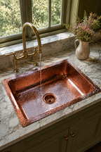 A hammered copper farmhouse sink with a matching polished brass bridge faucet, installed in a white marble countertop with green cabinets and a window view in the background.