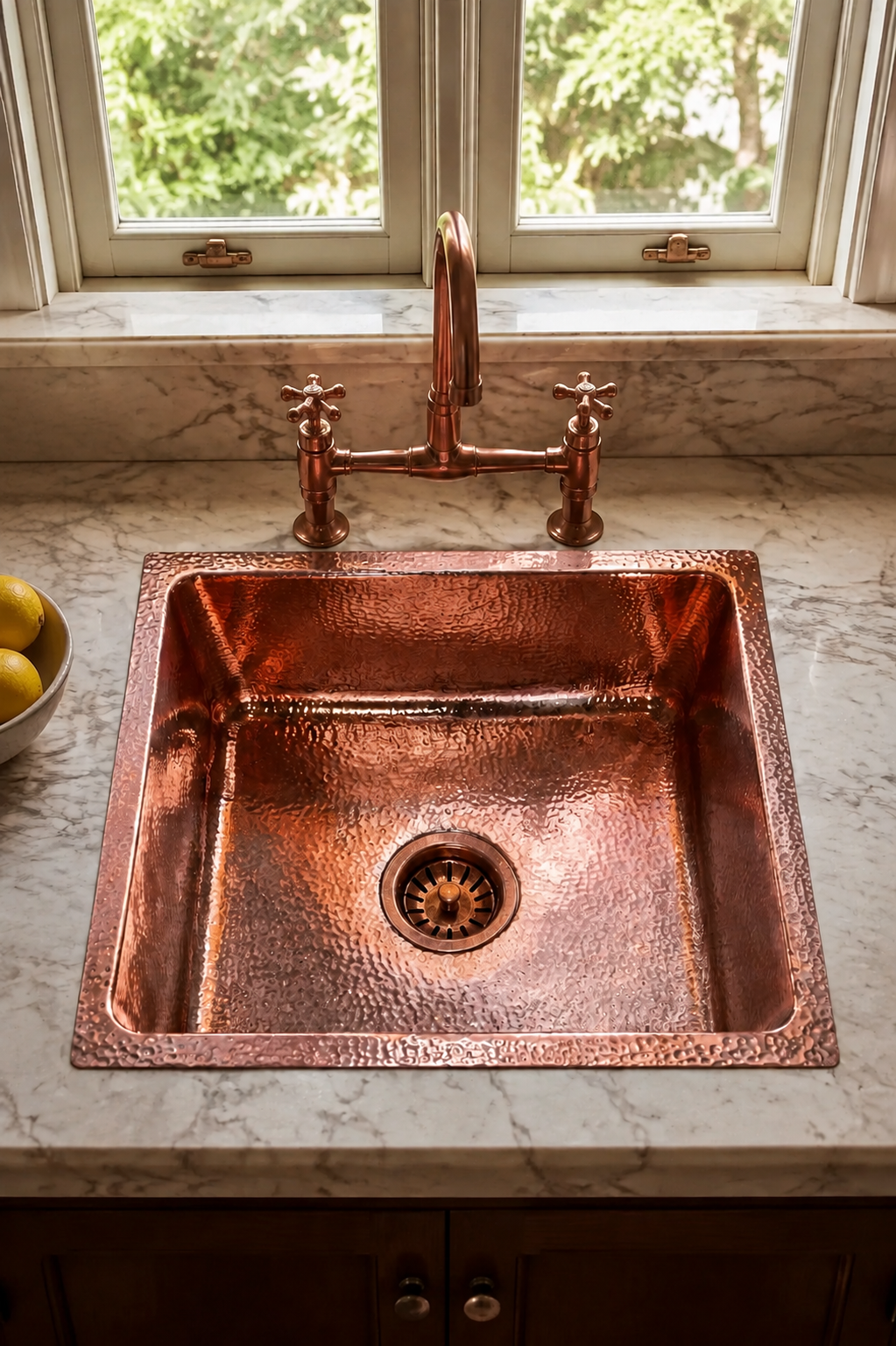 A hammered copper farmhouse sink with a matching copper bridge faucet and two cross-handle valves, installed in a white marble kitchen countertop beneath a window.