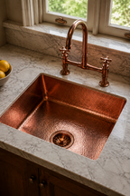 A hammered copper kitchen sink with a matching copper bridge faucet and two cross-handle valves, set into a light gray marble countertop near a window.