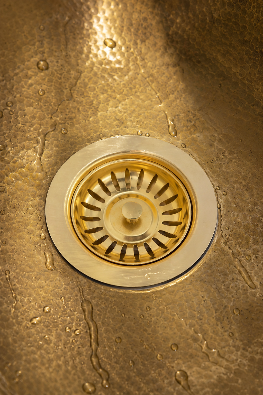 Close-up of a round metal kitchen sink drain set into a hammered metallic sink surface with water droplets visible.