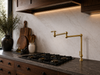 A modern kitchen scene featuring a polished brass pull-down faucet with an adjustable gooseneck spout, installed next to a black gas cooktop on a dark stone countertop. The background consists of a light beige marble backsplash, dark wood cabinetry, and decorative wooden cutting boards and a dark ceramic vase with dried branches.