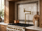 A close-up of a modern kitchen featuring a polished brass pull-down faucet, a white gas range with brass knobs, a copper pot, and a decorative tiled backsplash.