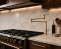 A close-up of a modern kitchen featuring a polished brass farmhouse-style faucet with a high-arc spout, set against a white marble backsplash and countertop with dark wood cabinetry.