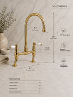 A polished brass bridge faucet with two white ceramic handles is shown on a white marble countertop with overlaid dimension lines and text detailing measurements and product specs.