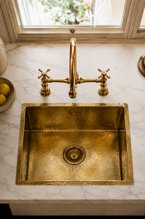 A hammered brass farmhouse sink with a matching bridge faucet and cross-handle valves, installed on a white marble countertop near a window.