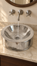 A hammered silver metal vessel sink with a waterfall faucet, installed on a beige marble bathroom countertop with dark wood cabinetry.