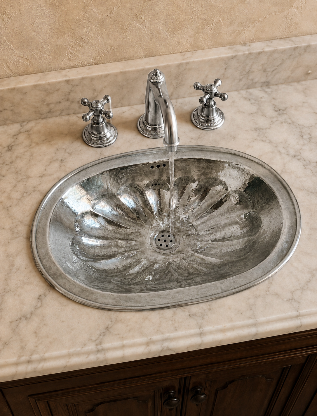 An oval silver metal sink basin with a decorative leaf-like pattern, installed in a beige marble countertop. A chrome faucet with water running into the sink is centered above it, with two matching chrome cross-handle knobs on either side.