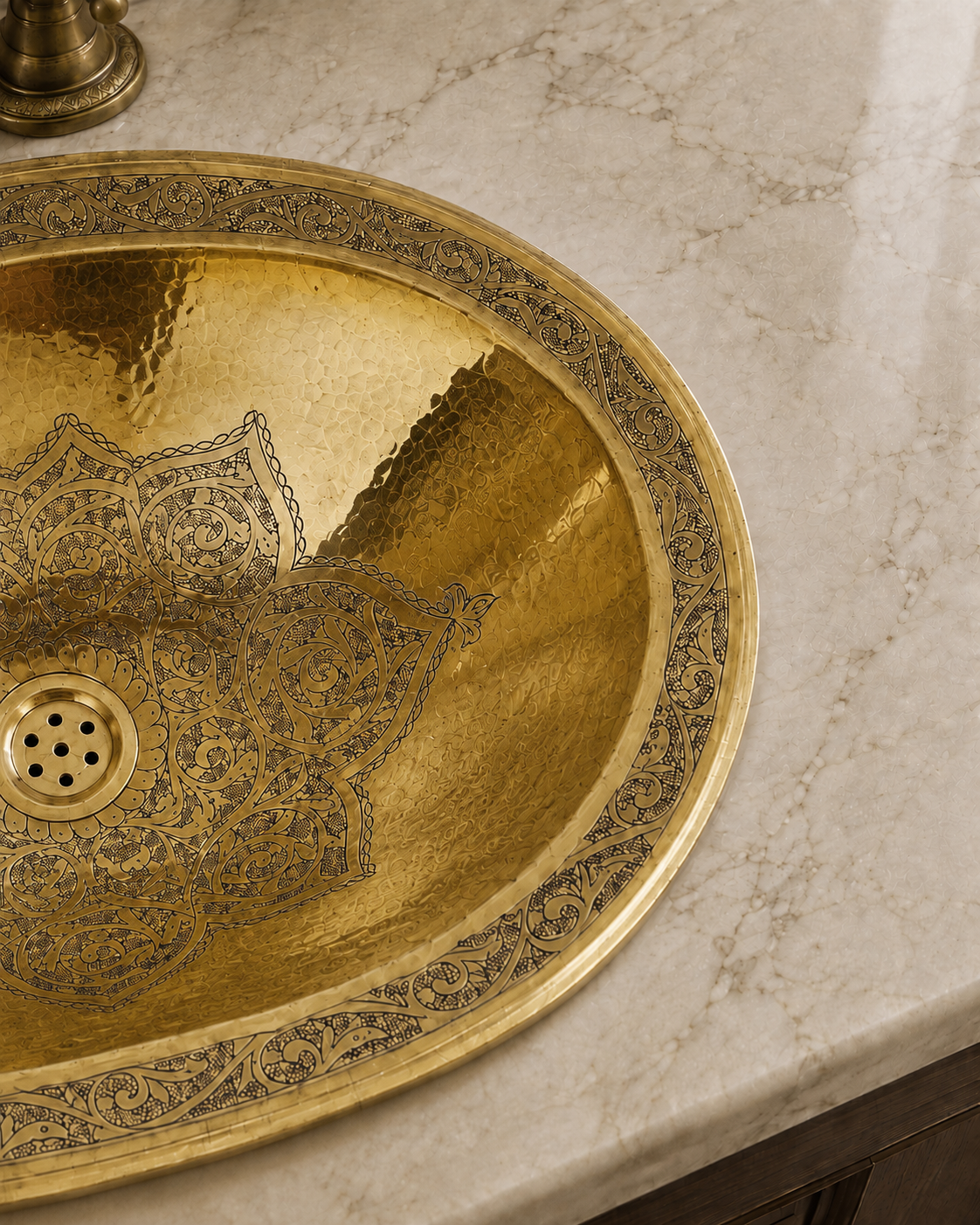 An oval hammered brass bathroom sink with intricate dark blue floral mandala engravings, set into a light beige marble countertop.